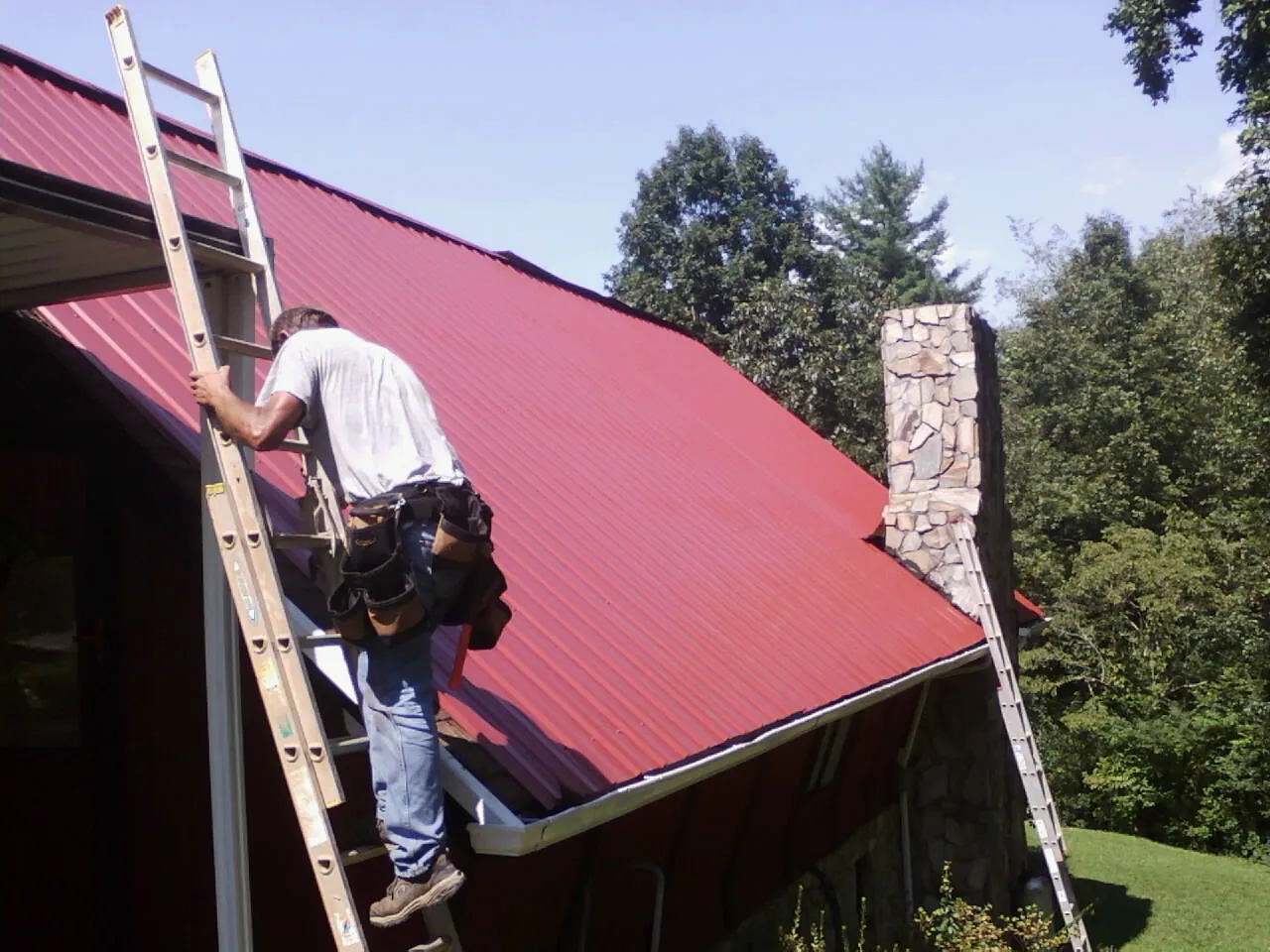 Roofer on a ladder beside a red metal roof and stone chimney — Soffit & Fascia Repair by Roofing Brothers in Ridley Park